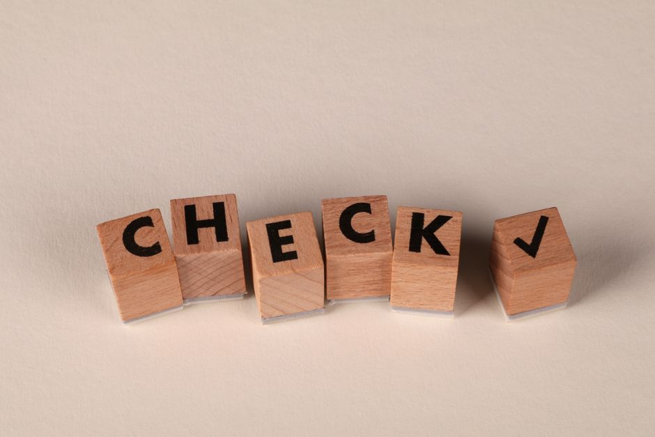 Six small wooden blocks with black uppercase letters spelling 'CHECK' are arranged in a straight line on a smooth, light-colored surface. The blocks have a natural wood finish with visible grain textu