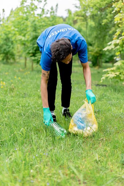A group of five people standing outdoors on a grassy field under a partly cloudy sky, engaged in a community clean-up or rubbish collection activity. From left to right, the first individual is an Afr