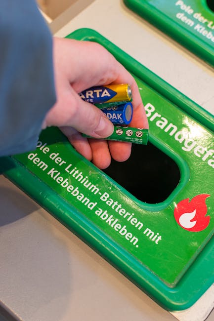 A person's hand holding a yellow and blue lithium battery over a green recycling bin designed for battery disposal, which features a circular opening in the centre. The person's hand is positioned abo
