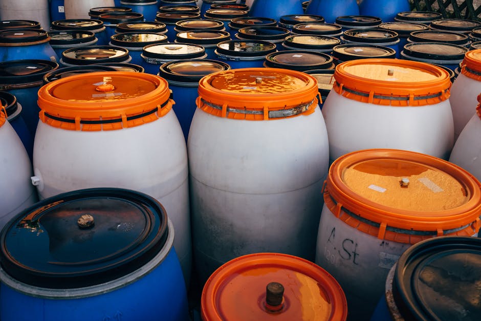 A large outdoor storage area filled with numerous plastic drums and barrels, primarily white with orange and black lids. The barrels are arranged in rows, with some standing upright and others lying o