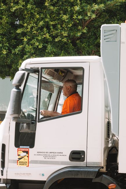 A waste collection worker wearing a black jacket with a high-visibility yellow vest, featuring reflective strips, operates a large, white refuse collection vehicle parked on an urban street during dus